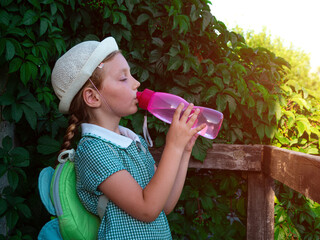 Cute school little girl drinks water from reusable pink bottle outdoor. Child in hat enjoys fresh...