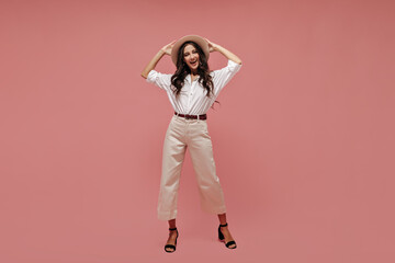 Photo of fashionable lady with curly hair in white shirt, light pants and black heels holding her wide-brimmed hat and smiling..