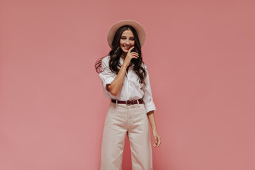 Lovely girl with long dark curly hair in cool light shirt, stylish trousers and white hat smiling...
