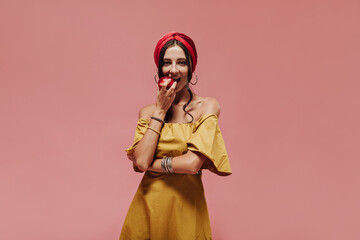 Modern woman in red headband, round earrings and yellow dress eating apple and looking into camera on isolated background..