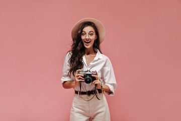 Cheerful curly lady with long hair in straw hat, white shirt and beige cool pants with belt looking into camera and holding camera..