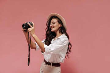 Lovely long wavy haired girl in modern eyeglasses, light wide blouse, beige pants with belt and hat smiling and posing with cool camera..