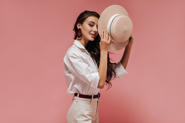 Beautiful lady with long wavy hair in modern brown earrings, light blouse and cool pants covers her eye with hat on pink backdrop..