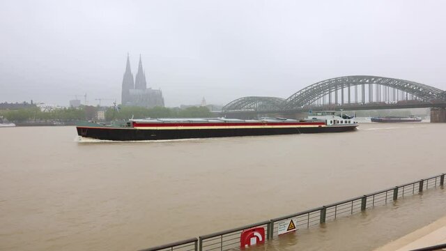 Koeln, Deutschland, 14.07.2021: Ein Frachtschiff faehrt durch das Hochwasser des Rheins unter der Hohenzollernbruecke vor dem Dom hinter dem ueberschwemmten Rheinboulevard in Koeln Deutz.