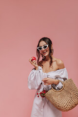 Smiling dark haired girl in white unusual sunglasses and lilac clothes holding apple and straw handbag on isolated background..