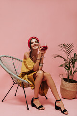 Smiling curly girl with bright headband, stylish dress and dark shoes holding glass with red wine and sitting on cool chair..
