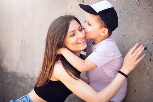 Little cute stylish boy hugs and kisses his mom on the cheek against the background of a gray concrete wall.