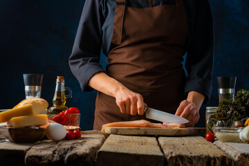 The chef prepares a Caesar salad. He cuts the chicken fillet into chunks on a cutting board. On a rough wooden table are the ingredients used in the salad. Dark blue background.