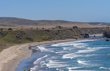 San Simeon, CA, USA - June 8, 2021: Pacific Ocean coastline North of town. Landscape with beige beach, green covered cliffs and white surf of deep blue ocean under light blue sky.