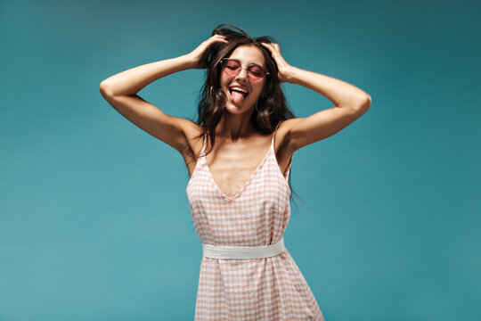 Funny stylish girl with curly dark hair in pink cpp sunglasses and plaid fashionable clothes showing her tongue with closed eyes..
