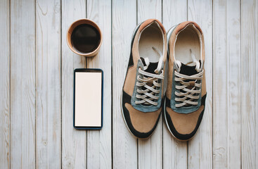 Stylish suede sneakers stand on a wooden gray background with hot coffee. Sport, morning, movement, copy space, fees for work. A pair of trend shoes, phone with white screen, mockup.