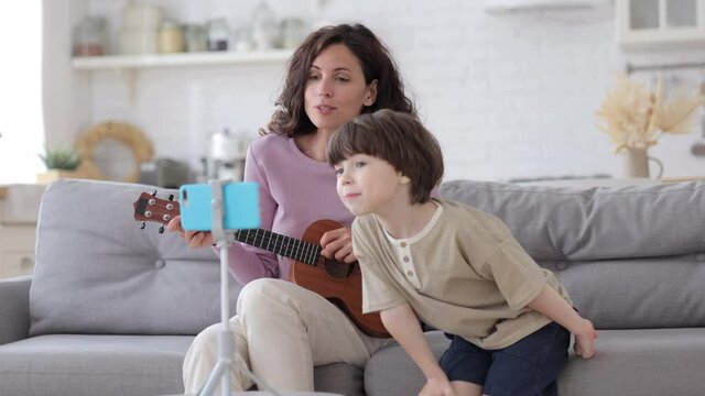Happy mother bonding with preschool kid play guitar, sing songs and record video on smartphone for relatives or dad. Working mom enjoy spending free time on quarantine at home with small son has fun