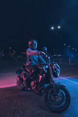 Motorcyclist sits on a motorbike in neon light in an empty parking lot at night - 445795407