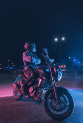 Naklejka na ściany i meble Motorcyclist sits on a motorbike in neon light in an empty parking lot at night