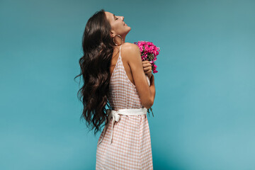Lovely long dark haired slim woman with tattoo in stylish sundress with wide white belt posing with pink flowers on blue background..