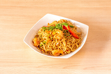 Yakisoba noodles with shrimp and peeled prawns, chopped chives and sauteed peppers and vegetables in white bowl and light wooden table