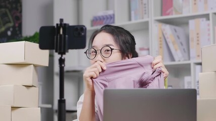 Happy young Asian woman wearing glasses, sitting and selling clothes by live broadcasting through internet channels at home, setting camera on a table,  online sales channel, woking at home