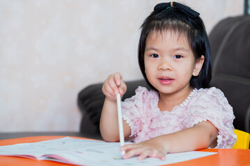 Adorable child girl holding pencil and her pointing it on the work book. Kid doing homework. Children looking at the camera. Lady sweet smiling. Baby have a question.