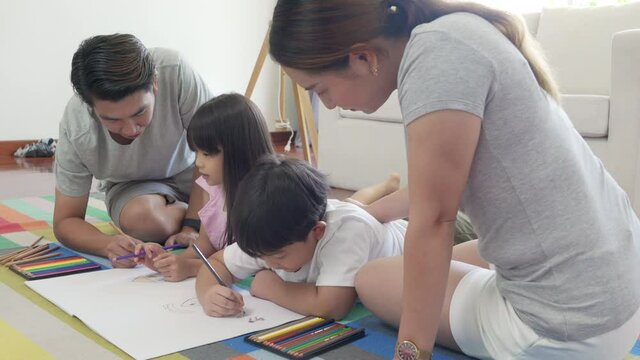 Asian family parents with little daughter and son lying on the floor using color pencil drawing on paper book together. Father and mother with cute boy and girl kid having fun homeschooling at home