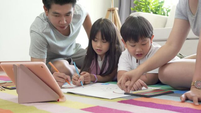 Asian family parents with little daughter and son lying on the floor using color pencil drawing on paper book together. Father and mother with cute boy and girl kid having fun homeschooling at home