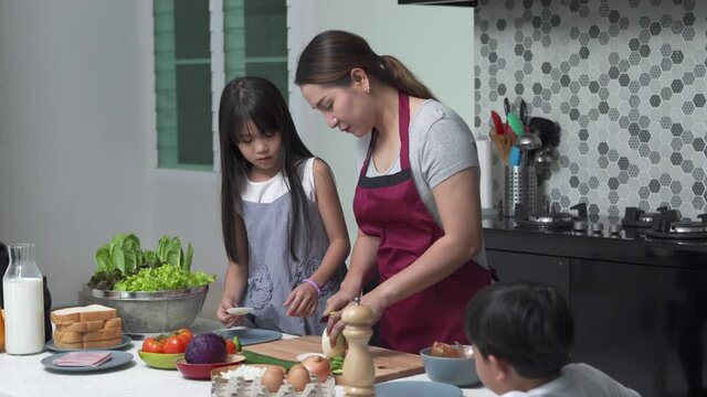 Asian mother teach little daughter and son making vegetable and ham sandwich on the table for breakfast in kitchen. Mom and child boy and girl kids enjoy weekend activity cooking together at home.