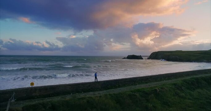 Aerial: Man walking along ocean wall on stormy day with rough ocean. Annestown Beach, Tramore, Ireland