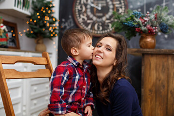 Fototapeta na wymiar Little boy is kissing his mother near fir-tree. Christmas celebration. Son with Mom weared in red checkered shirts. New year eve