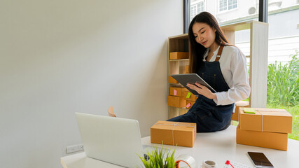 Independent Asian woman SME business owner online shopping working on laptop computer with parcel box on table at home - online SME business and online shipping and shopping concept.