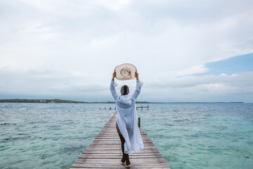 Woman tourist in white dress holding hat and walking on the wooden dock enjoying summer holiday