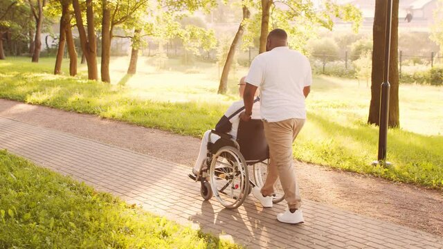 African-American caregiver and old disabled man in a wheelchair. Professional nurse and handicapped patient in the park. Assistance, rehabilitation and health care. Golden hour sunset.