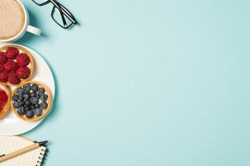 Top view photo of workplace glasses cup of frothy coffee pen notebook and plate with three berry cupcakes on isolated pastel blue background with empty space