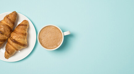 Top view photo of plate with two fresh croissants and cup of frothy coffee on isolated light blue background with empty space