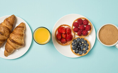 Top view photo of two plates with three berry cupcakes and two croissants glass of juice and cup of frothy coffee on isolated light blue background
