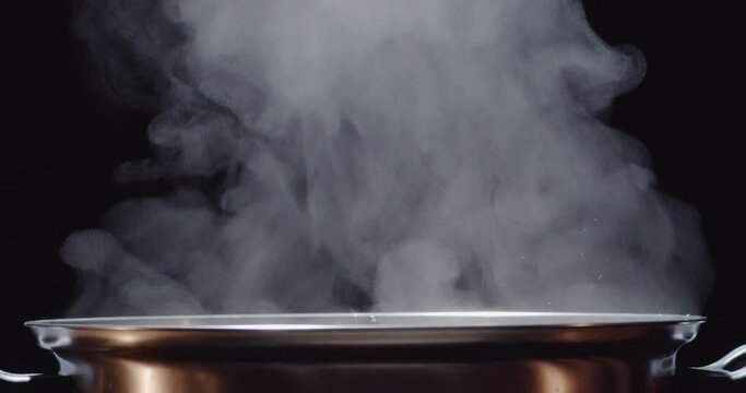 Slow motion of hot pot or shabu-shabu pot made of stainless steel, water boiling, hot steam rising on a black screen, a hand put vegetable into the pot.