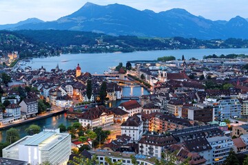 Night scenery of beautiful Lucerne by lakeside with landmark Chapel bridge (Kapellbrucke) across Reuss River, majestic Rigi mountain in background and city lights glowing at blue dusk, Switzerland