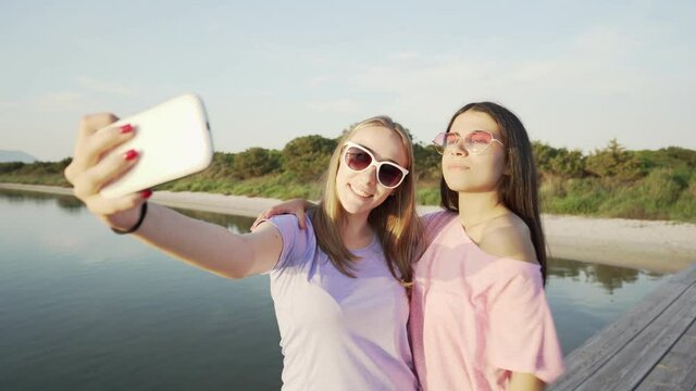 Young women friends couple with colored sunglasses making silly faces taking selfie with smartphone at sunset or dawn at sea or lake resort. Blonde girl having fun sharing vacation on social network