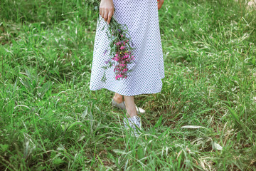 the legs of a beautiful girl in a long white dress and gray shoes are standing in the tall green grass. A girl holds a bouquet of purple wildflowers. Selective focus. Copy Space