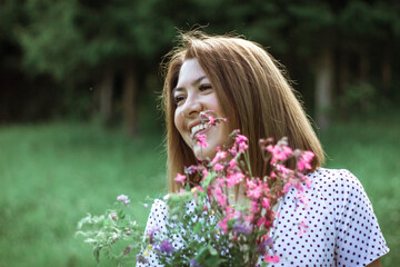 Fototapeta premium A girl with a short haircut and flowers in her hands looks away and laughs. A close portrait of a young beautiful woman in a meadow with lilac wildflowers in her hands. Selective focus. Copy space