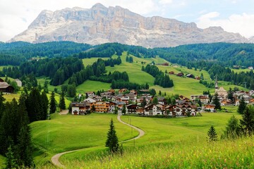 Fototapeta na wymiar Summer scenery of a beautiful village at foothills of rugged Sasso di Santa Croce & a country road winding in the green grassy valley in Val Badia, Alta Badia, Dolomiti, Trentino, South Tyrol, Italy