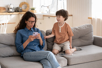 Indifferent young mother surfing social media addict irritated with disturbing little son. Mom...