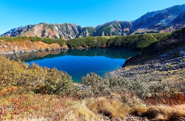 Fall scenery of beautiful Mikurigaike Pond, a crater lake in Murodo Tateyama, Toyama, Japan, with majestic mountain in background under clear blue sunny sky on a brisk autumn day ~A hiking destination