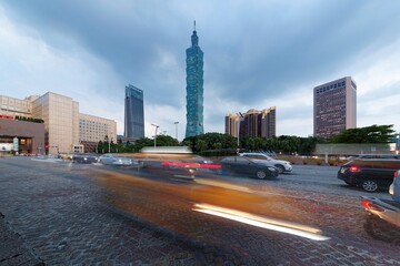 Scenery of a busy street corner at rush hour in Downtown Taipei, capital city of Taiwan, with light trails of cars dashing by the City Hall & Taipei 101 Tower in Xinyi District under moody cloudy sky