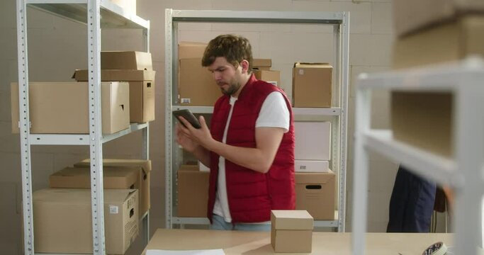 Man in red vest, employee of warehouse putting parcels on table and typing on gadget tablet, checking information about delivery box on the background of cardboard boxes. Logistics, storage concept. 
