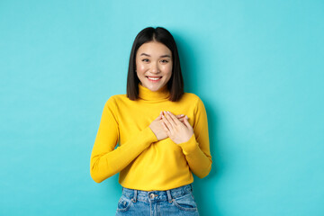 Image of beautiful asian woman holding hands on heart and smiling, thanking you, feeling grateful, standing over blue background