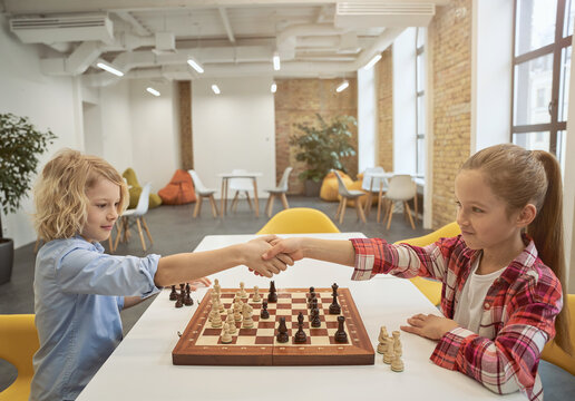 Smart little children, boy and girl shaking hands after match, playing board game, sitting together at the table in school
