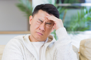 headshot of a young asian man holding his head with a severe headache at home in a room on the couch. Up close, the casual male massages the temples and suffers from chronic pain and fatigue. Closeup