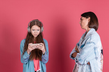 Modern mom and daughter in denim jackets on terracotta background girl in headphones play game on phone and disappointed angry mom stands by