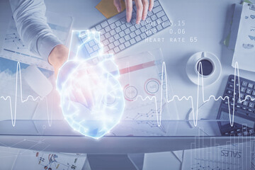 Double exposure of man's hands typing over computer keyboard and human heart hologram drawing. Top view. Medical education concept.