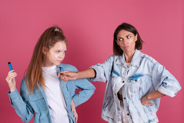 Modern mom and daughter in denim jackets on terracotta background, kid smokes electronic cigarette, angry mom stands by