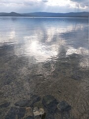 The sun's rays break through the clouds and are reflected in a transparent lake at the bottom of which large sand and sharp stones are visible